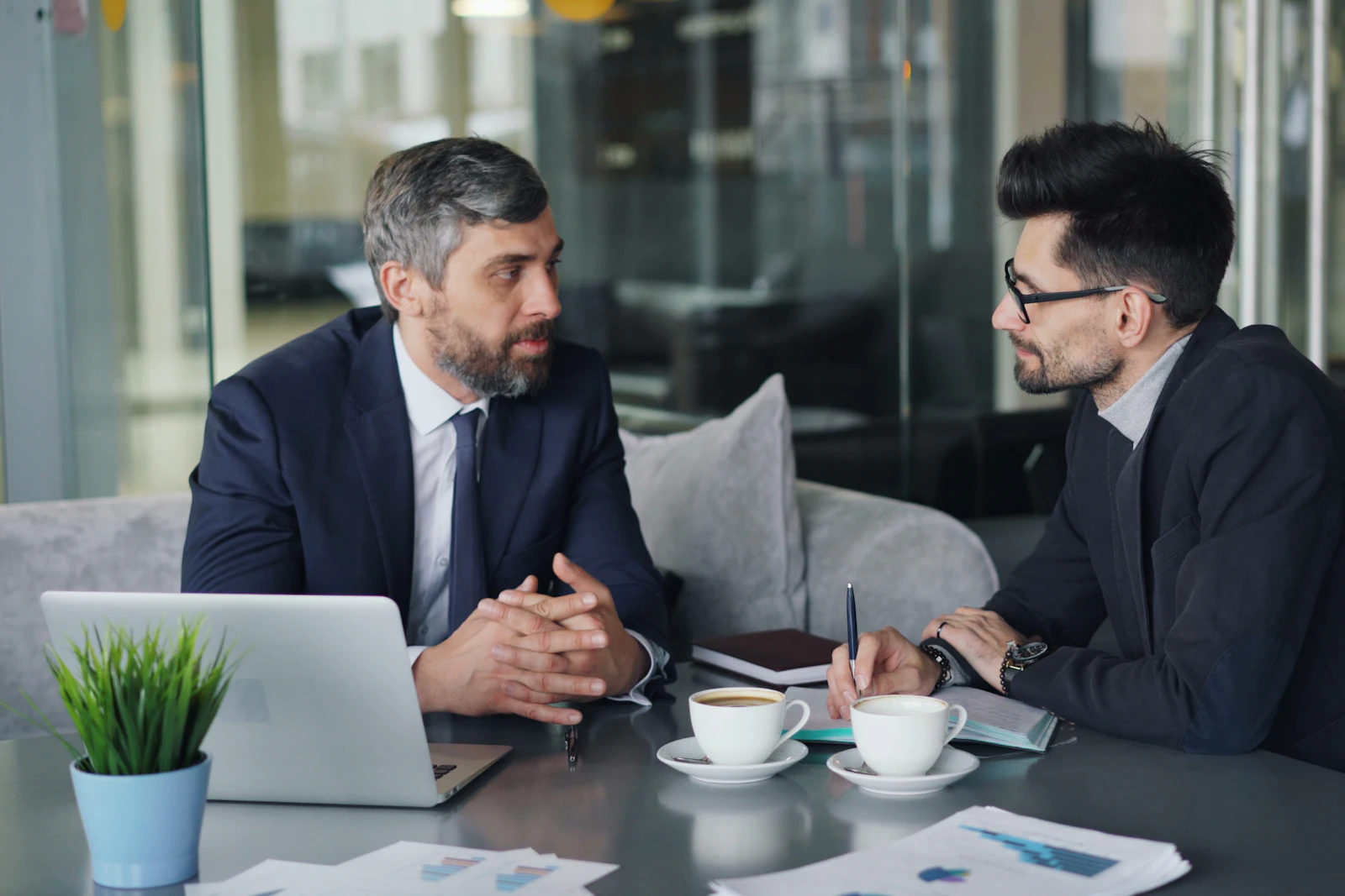 Twee mannen in gesprek aan een tafel met koffie, een laptop en uitgeprinte grafieken