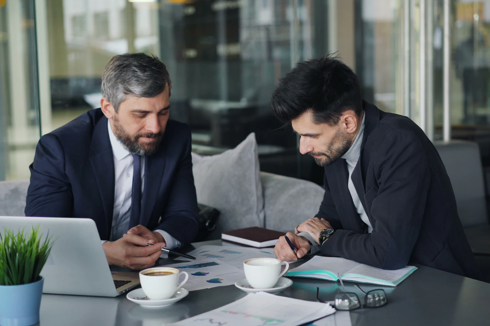 Twee mannen in pak bekijken uitgeprinte grafieken aan een tafel, met een laptop en koffie