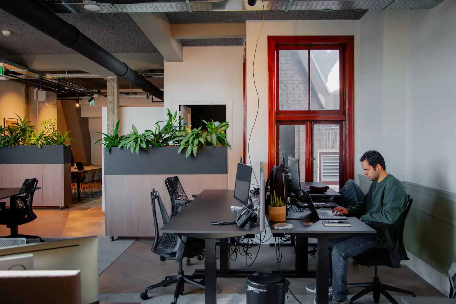 Man werkt achter een laptop in een licht kantoor met planten en een rood kozijn