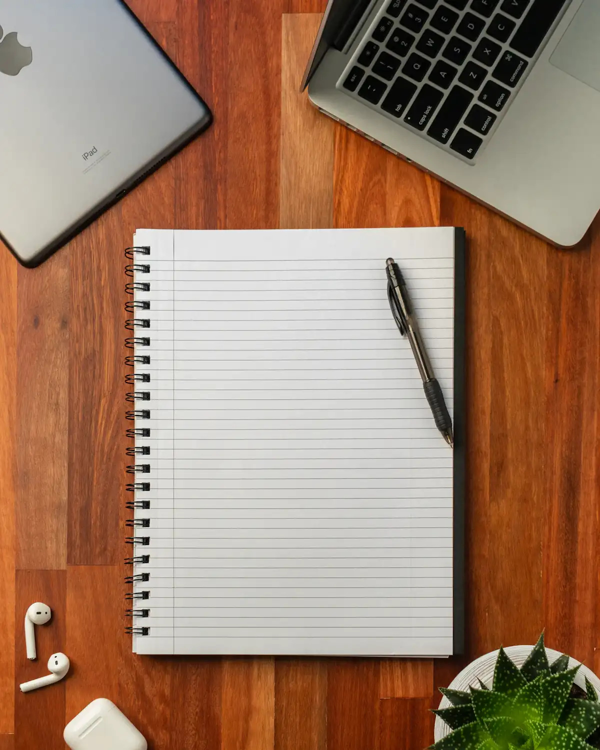 Leeg notitieblok met pen op een houten bureau, met een laptop en een plant eromheen