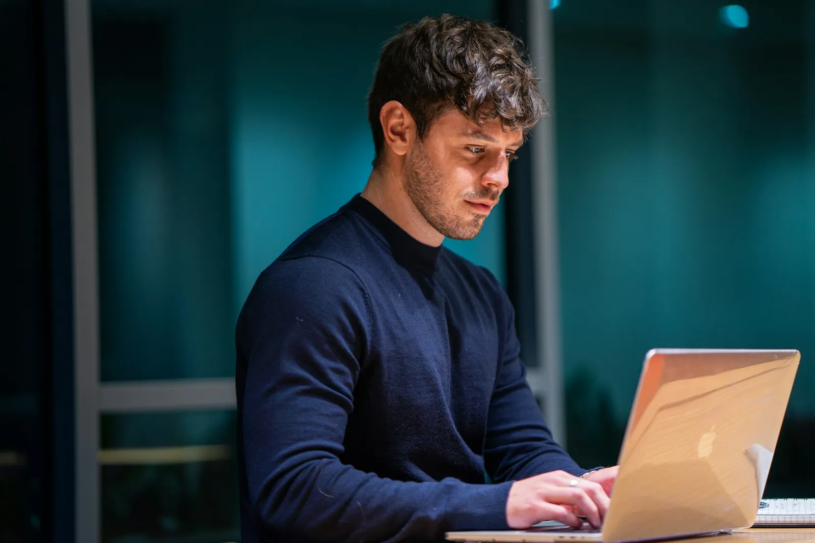 Man in een donkerblauwe trui werkt geconcentreerd op een laptop aan een houten tafel