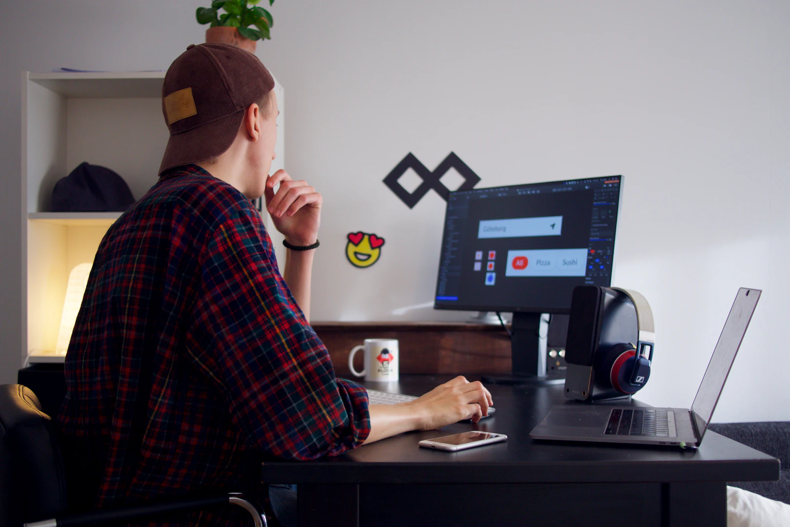 Man met pet aan een bureau met een beeldscherm en een laptop
